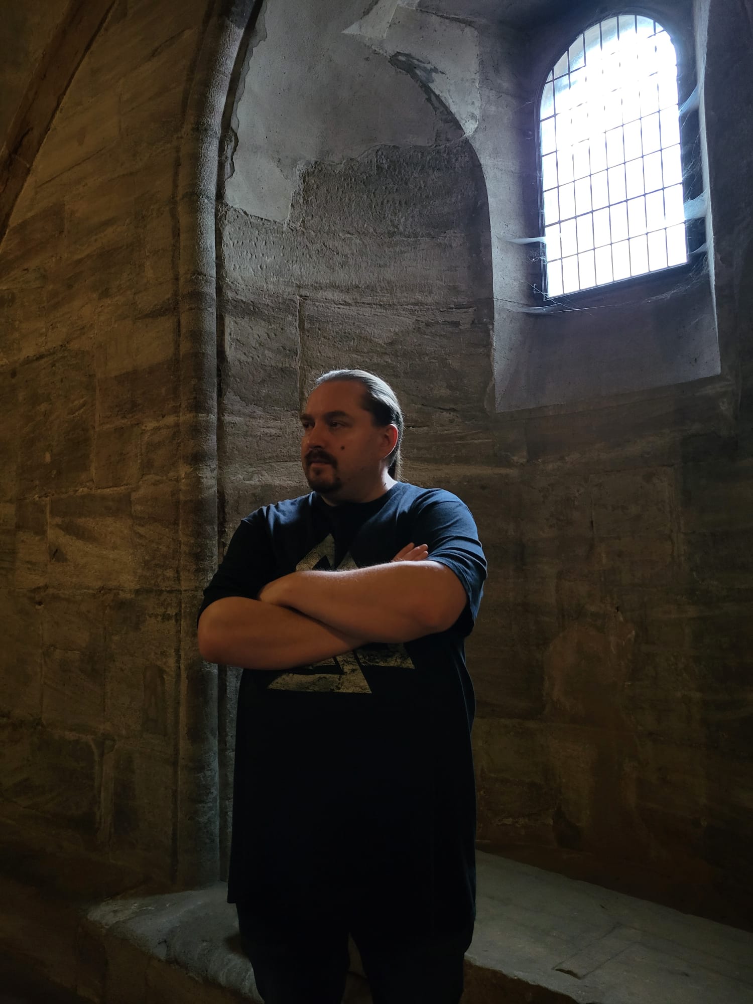 Bane in a stone chamber, standing with folded arms beneath a tall arched window.