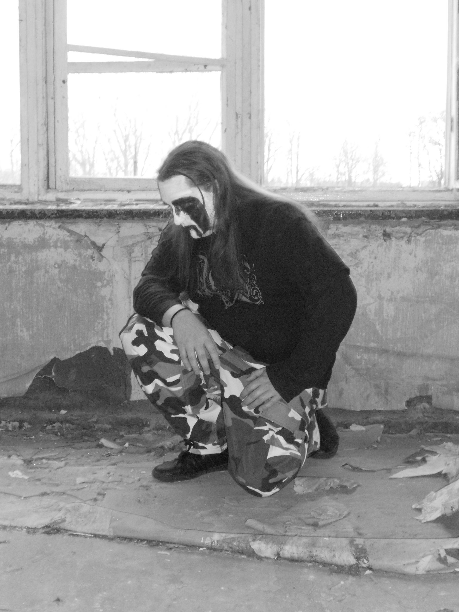 Early black and white photo of Bane with corpsepaint, kneeling in a ruined room.